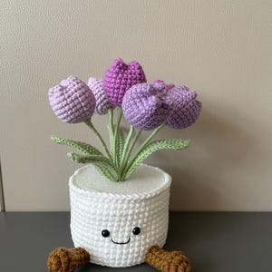 May include: A handmade crochet flower pot with a smiling face and brown feet. The pot is white and holds several purple and lavender crochet tulips with green stems and leaves. A charming, whimsical decorative item.