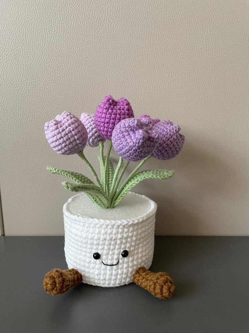 May include: A handmade crochet flower pot with a smiling face and brown feet. The pot is white and holds several purple and lavender crochet tulips with green stems and leaves. A charming, whimsical decorative item.