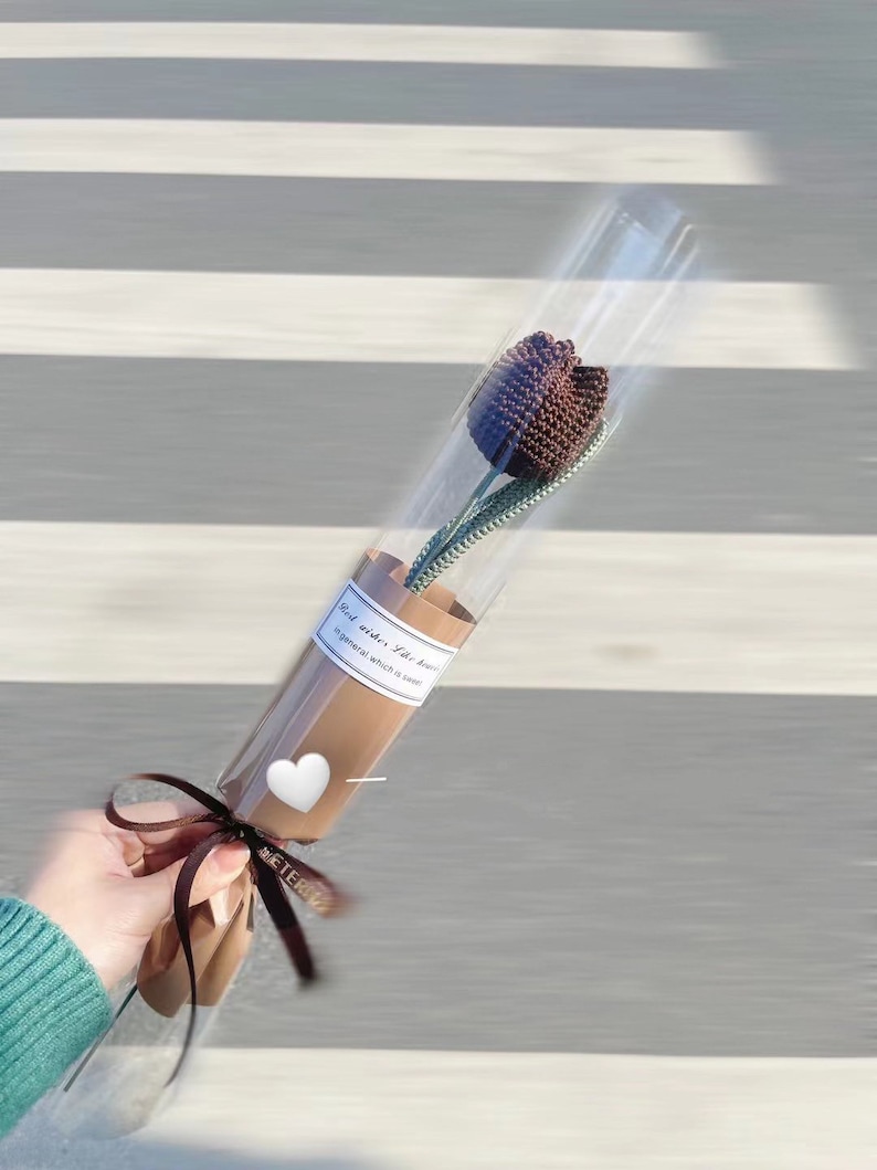 K&ouml;nnte beinhalten: Eine einzelne, dunkelbraune geh&auml;kelte Tulpenblume in einer transparenten Plastikh&uuml;lle, gebunden mit einem braunen Band. Der Stiel ist gr&uuml;n. Ein wei&szlig;es Etikett mit Text und ein wei&szlig;er Herzaufkleber sind sichtbar.