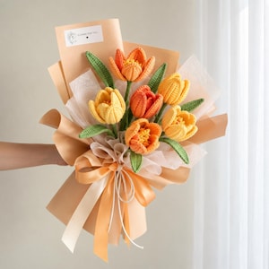 May include: A bouquet of crocheted tulips in shades of yellow and orange, featuring green crocheted leaves and stems. The flowers are wrapped in peach-colored paper with a matching satin ribbon bow and a small white tag labeled Flower For You.