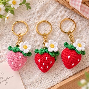 Pode incluir: Três porta-chaves de morango em croché com argolas douradas. Um é rosa claro e dois são vermelhos, cada um com sementes brancas, folhas verdes e uma pequena flor branca com centro amarelo. Um cartão diz: Small Things Bring Big Happiness.