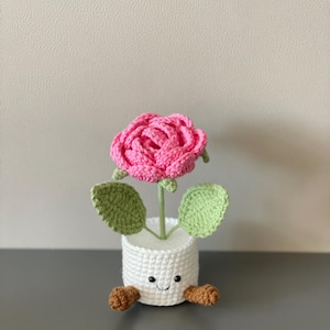 May include: A handmade crochet flower in a white pot with a smiling face and brown feet. The pink rose has green leaves and a green stem. This whimsical, decorative item is perfect for home decor.