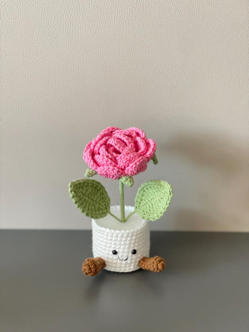 May include: A handmade crochet flower in a white pot with a smiling face and brown feet. The pink rose has green leaves and a green stem. This whimsical, decorative item is perfect for home decor.