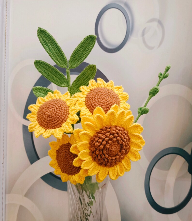 May include: A bouquet of crocheted sunflowers in a clear glass vase. The sunflowers are yellow with brown centers and green leaves.