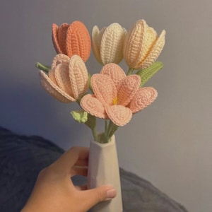 May include: A bouquet of crocheted tulips in shades of pink and cream, arranged in a white ceramic vase.