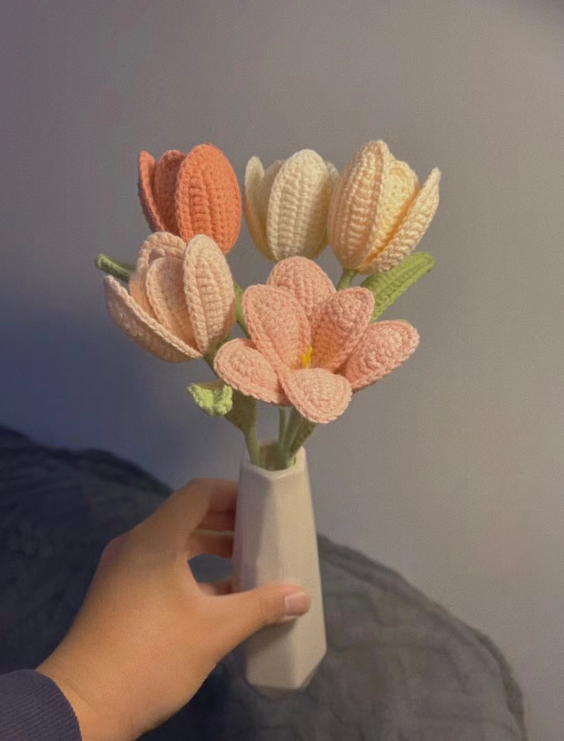 May include: A bouquet of crocheted tulips in shades of pink and cream, arranged in a white ceramic vase.