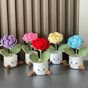 May include: A collection of six crocheted flower arrangements. Each flower is a different color: purple, pink, red, yellow, and blue. The flowers sit in white pots with smiling faces and brown feet, and green leaves.