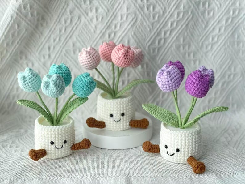 May include: Three crochet tulip arrangements in white pots with smiling faces and brown arms and legs. The tulips are in shades of blue, pink, and purple. The pots are on a white surface.