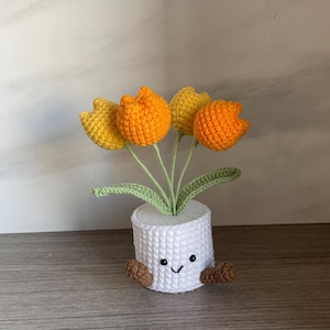 May include: A crocheted flower pot with a smiling face, brown feet, and five orange and yellow crocheted tulips. The white pot has a textured, woven appearance, and the green leaves add a natural touch.