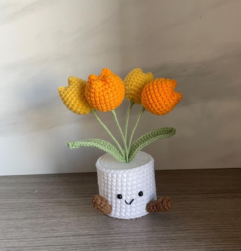 May include: A crocheted flower pot with a smiling face, brown feet, and five orange and yellow crocheted tulips. The white pot has a textured, woven appearance, and the green leaves add a natural touch.