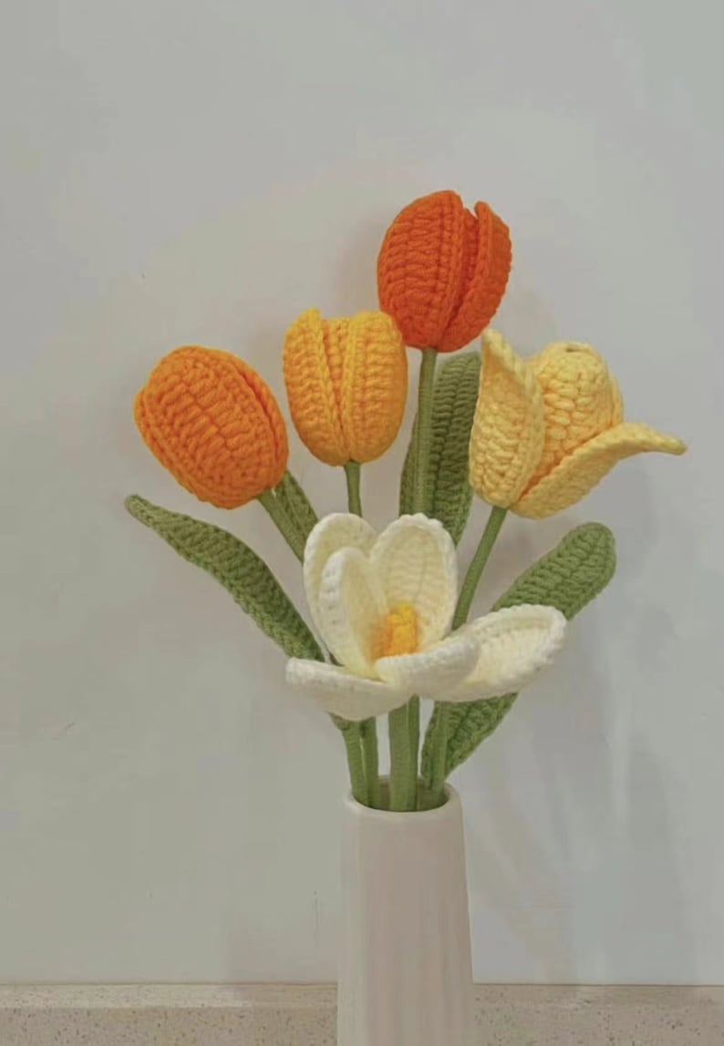 May include: A bouquet of crocheted flowers in a white vase. The flowers are orange, yellow, and white. The flowers are arranged in a vase with a white background.