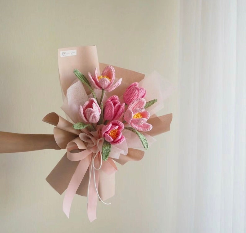 May include: A bouquet of pink crocheted flowers with green leaves wrapped in pink tissue paper and tied with a white ribbon.