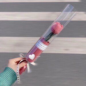 K&ouml;nnte beinhalten: Eine rosa geh&auml;kelte Tulpenblume in einer transparenten Plastikr&ouml;hre mit rotem Sockel. Die R&ouml;hre ist mit einem wei&szlig;en Band und einem herzf&ouml;rmigen Aufkleber gebunden. Der Text auf dem Etikett lautet "Best wishes for..."