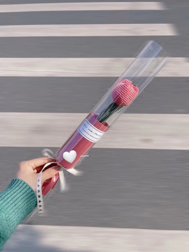 K&ouml;nnte beinhalten: Eine rosa geh&auml;kelte Tulpenblume in einer transparenten Plastikr&ouml;hre mit rotem Sockel. Die R&ouml;hre ist mit einem wei&szlig;en Band und einem herzf&ouml;rmigen Aufkleber gebunden. Der Text auf dem Etikett lautet "Best wishes for..."