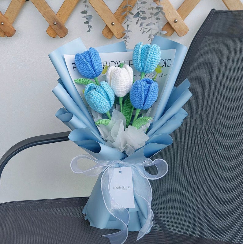 May include: A bouquet of crocheted blue and white tulips wrapped in light blue tissue paper with a white ribbon and a tag that says "Seeds & Blooms".