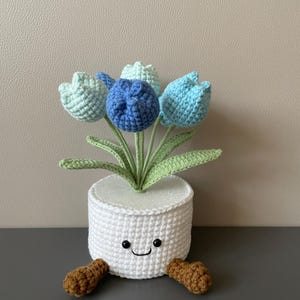 May include: A crocheted white pot with a smiling face holds a bouquet of blue and green tulips. The pot has brown feet. The flowers are in shades of blue and green, with green leaves. A charming handmade decorative item.