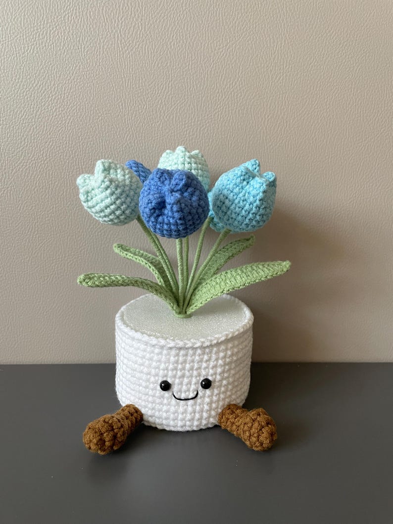 May include: A crocheted white pot with a smiling face holds a bouquet of blue and green tulips. The pot has brown feet. The flowers are in shades of blue and green, with green leaves. A charming handmade decorative item.