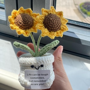 May include: Crocheted sunflower arrangement in a white pot with a smiling face. The sunflowers have yellow petals and brown centers. The pot has a small white tag with the text: "Life can be tough sometimes, but remember so are you!"
