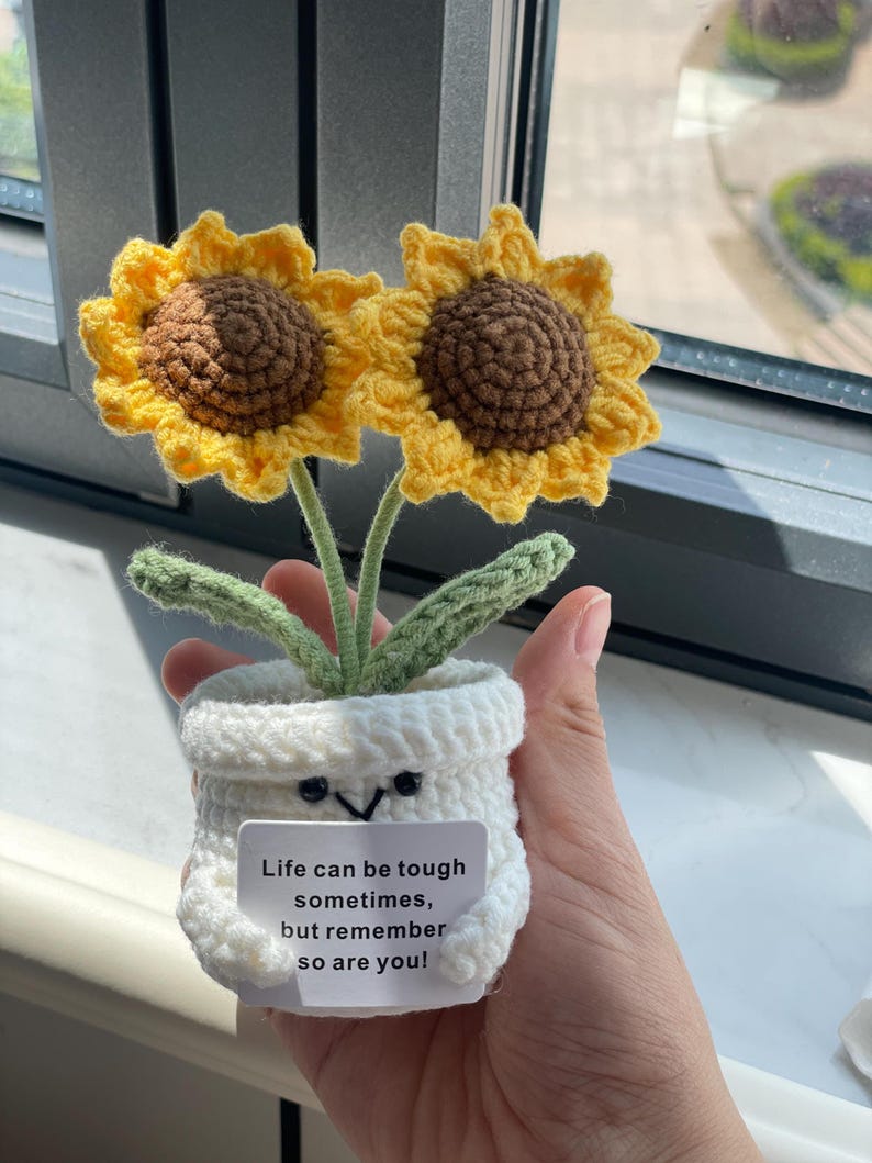 May include: Crocheted sunflower arrangement in a white pot with a smiling face. The sunflowers have yellow petals and brown centers. The pot has a small white tag with the text: "Life can be tough sometimes, but remember so are you!"