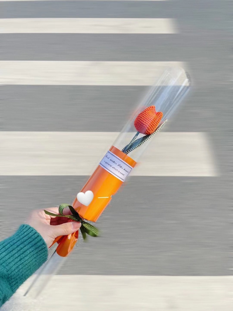 K&ouml;nnte beinhalten: Eine einzelne orangefarbene H&auml;keltulpe in einer transparenten Plastikh&uuml;lle, verpackt in orangefarbenem Papier und mit einem gr&uuml;nen Band gebunden. Die H&uuml;lle hat ein Etikett mit Text. Ein wei&szlig;er Herzaufkleber befindet sich auf dem orangefarbenen Papier.