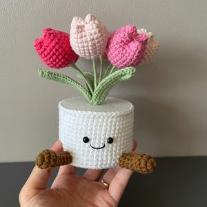 May include: A handmade crochet tulip arrangement in a white pot. The tulips are in shades of pink and the leaves are green. The white pot has a smiling face and brown feet.