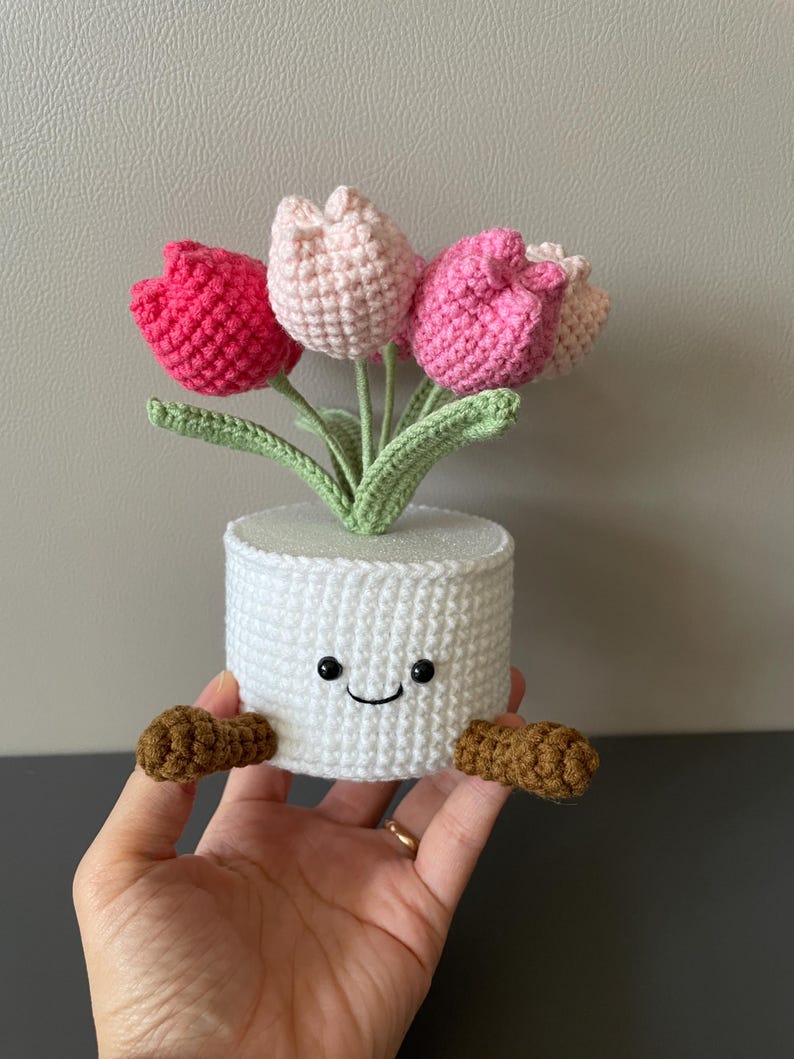 May include: A handmade crochet tulip arrangement in a white pot. The tulips are in shades of pink and the leaves are green. The white pot has a smiling face and brown feet.