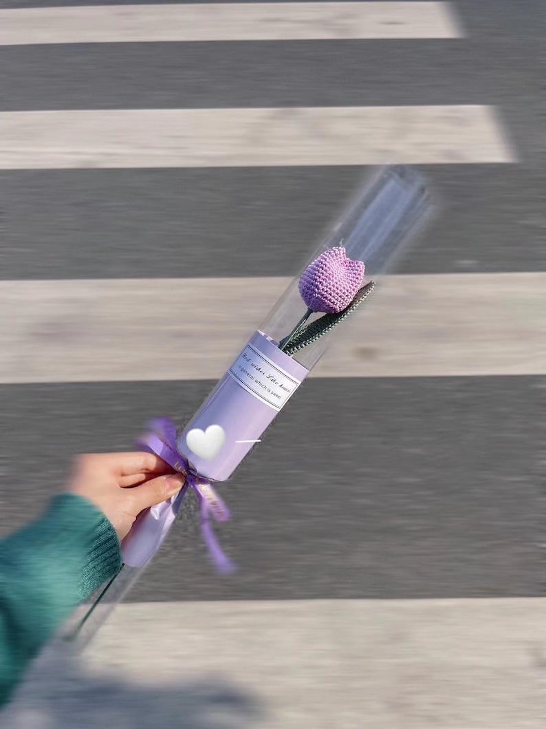 K&ouml;nnte beinhalten: Eine einzelne, handgefertigte, lavendelfarbene H&auml;keltulpe in einer transparenten Zellophanverpackung. Der Stiel ist gr&uuml;n, und die Blume ist mit einem lavendelfarbenen Band und einem wei&szlig;en herzf&ouml;rmigen Aufkleber gebunden. Die Verpackung tr&auml;gt den Text "a general which is seen".