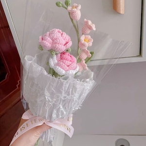 May include: A bouquet of crocheted flowers in pink and white, wrapped in clear plastic with a pink ribbon that says "Love is eternal".