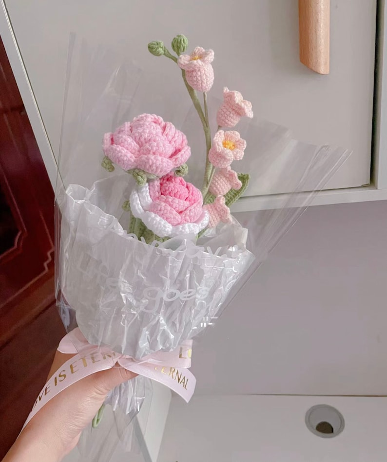 May include: A bouquet of crocheted flowers in pink and white, wrapped in clear plastic with a pink ribbon that says "Love is eternal".