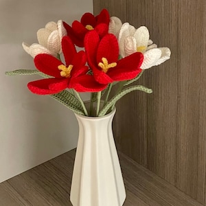 May include: A white ceramic vase with a textured surface holds a bouquet of crocheted flowers. The flowers are red and white, with yellow centers. The vase is on a wooden surface.