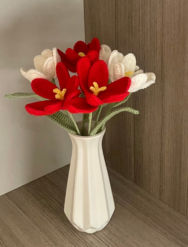 May include: A white ceramic vase with a textured surface holds a bouquet of crocheted flowers. The flowers are red and white, with yellow centers. The vase is on a wooden surface.