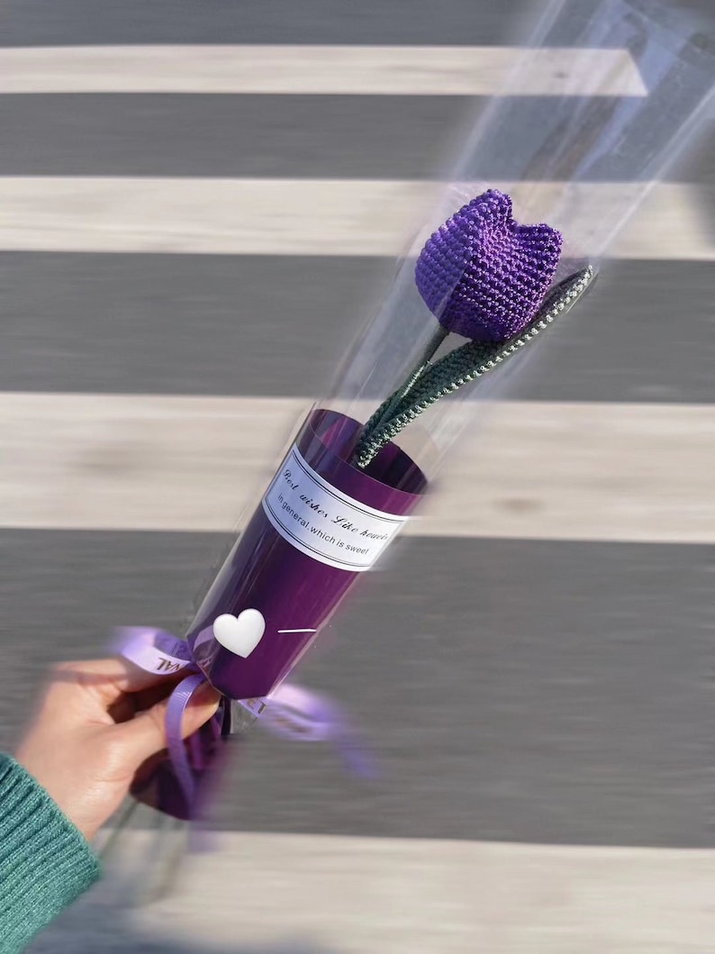 K&ouml;nnte beinhalten: Eine einzelne, handgefertigte lila H&auml;keltulpe in einem dekorativen lila Papierkegel. Der Kegel ist mit einer transparenten Plastikh&uuml;lle umwickelt und mit einem lila Band gebunden. Der Text auf dem Kegel lautet "Best wishes like heaven in general, which is sweet."