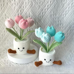 May include: Two crochet tulip flower arrangements in white pots with smiling faces and brown feet. One arrangement has pink tulips, the other has blue and teal tulips. The flowers have green stems and leaves.
