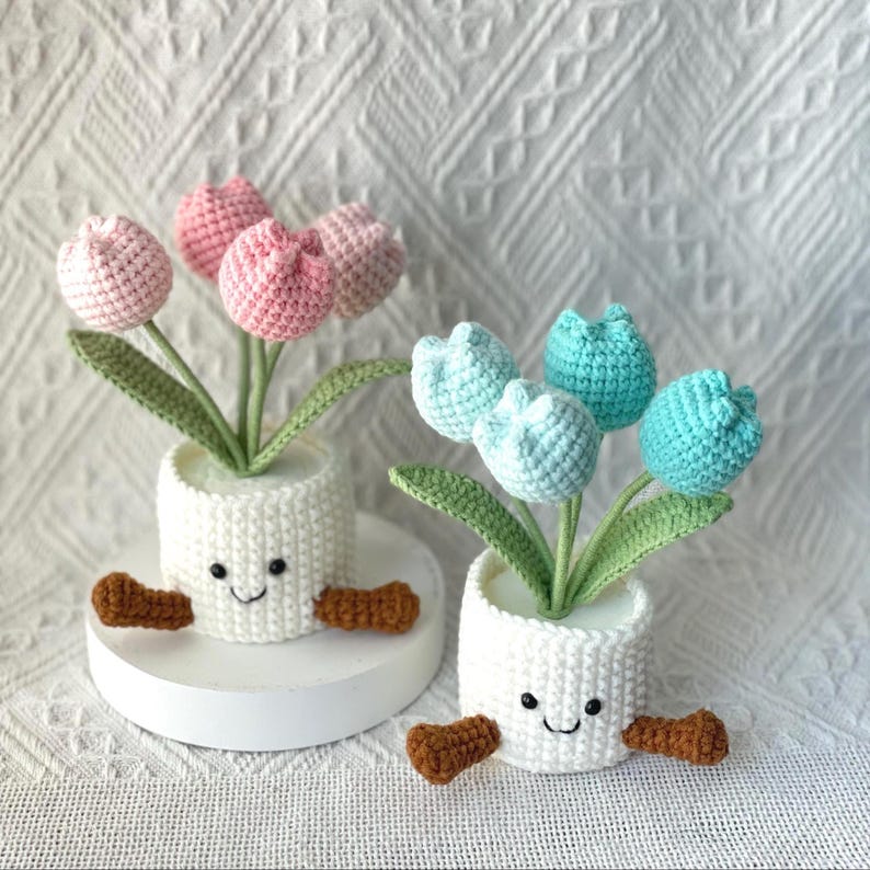 May include: Two crochet tulip flower arrangements in white pots with smiling faces and brown feet. One arrangement has pink tulips, the other has blue and teal tulips. The flowers have green stems and leaves.