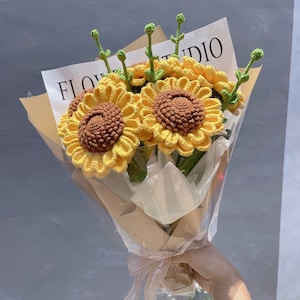 May include: A bouquet of crocheted sunflowers in a brown paper wrapper with a white ribbon tied around it. The bouquet is labeled "FLOWER STUDIO".