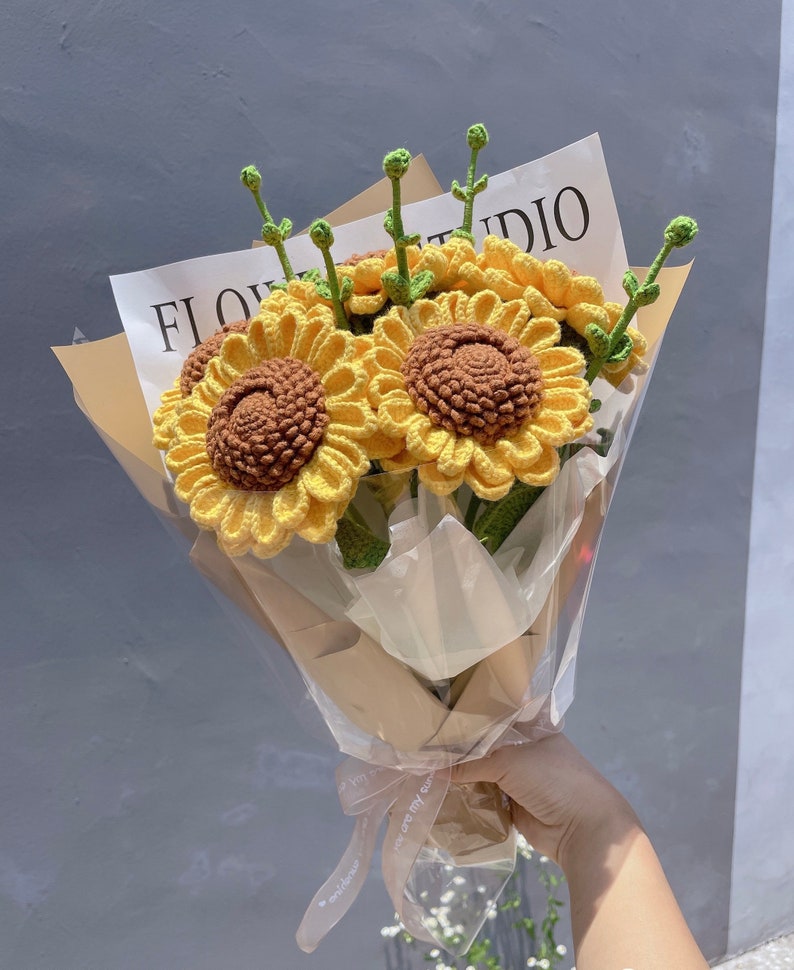 May include: A bouquet of crocheted sunflowers in a brown paper wrapper with a white ribbon tied around it. The bouquet is labeled "FLOWER STUDIO".