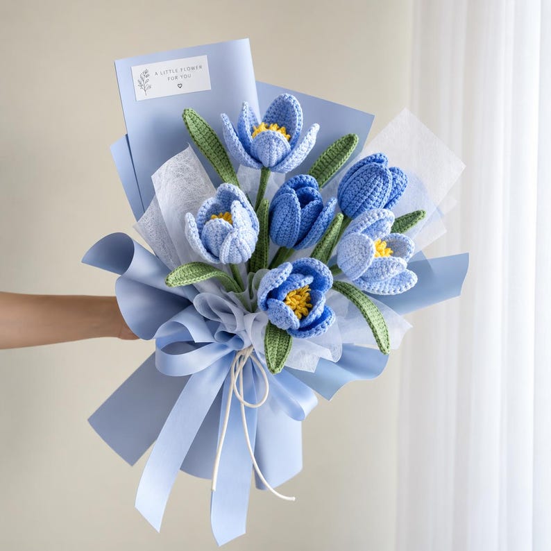 May include: A bouquet of crocheted blue tulips with yellow centers and green leaves, wrapped in light blue paper and white tissue paper. A small white tag at the top reads A LITTLE FLOWER FOR YOU with a small heart icon and a floral graphic.