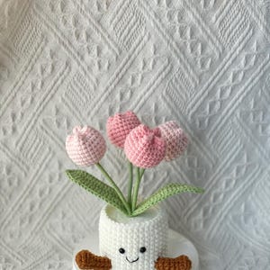 May include: A handmade crochet flower arrangement featuring pink tulips in a white pot with a smiling face and brown arms. The pot sits on a white circular base, creating a charming and whimsical decorative piece.