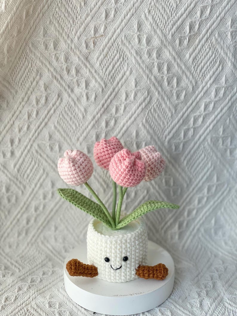 May include: A handmade crochet flower arrangement featuring pink tulips in a white pot with a smiling face and brown arms. The pot sits on a white circular base, creating a charming and whimsical decorative piece.