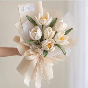 May include: A bouquet of six crocheted cream-colored tulips with yellow centers and green leaves, wrapped in beige paper with a matching ribbon. A small white rectangular tag at the top features the printed text A LITTLE FLOWER FOR YOU.