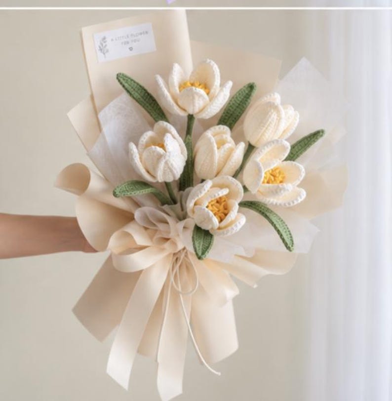 May include: A bouquet of six crocheted cream-colored tulips with yellow centers and green leaves, wrapped in beige paper with a matching ribbon. A small white rectangular tag at the top features the printed text A LITTLE FLOWER FOR YOU.