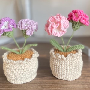 Op de afbeelding: Twee gehaakte bloemstukken in witte potten. Eén arrangement heeft roze bloemen en de andere heeft paarse bloemen.