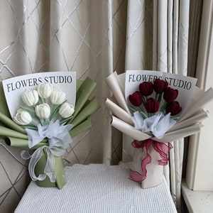 K&ouml;nnte beinhalten: Zwei Str&auml;u&szlig;e geh&auml;kelter Tulpen, einer wei&szlig; und einer rot, in Papier verpackt mit einem Band und dem Text "FLOWERS STUDIO".