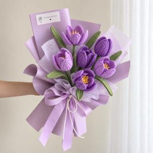 May include: A bouquet of six crocheted purple tulips with yellow centers and green leaves, wrapped in light purple paper with a white ribbon. A small white rectangular tag at the top features a line drawing of a flower and the text FLOWER FOR YOU.