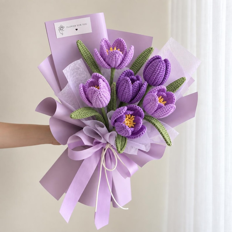May include: A bouquet of six crocheted purple tulips with yellow centers and green leaves, wrapped in light purple paper with a white ribbon. A small white rectangular tag at the top features a line drawing of a flower and the text FLOWER FOR YOU.