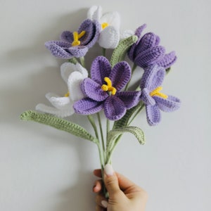 May include: A bouquet of crocheted purple and white flowers with green stems. The flowers are arranged in a hand.
