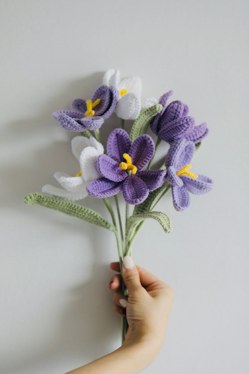 May include: A bouquet of crocheted purple and white flowers with green stems. The flowers are arranged in a hand.