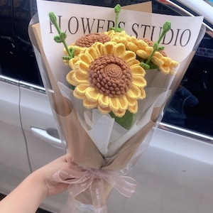 May include: A bouquet of crocheted sunflowers in a brown paper wrapper with a white ribbon tied around it. The bouquet is labeled "FLOWERS STUDIO".