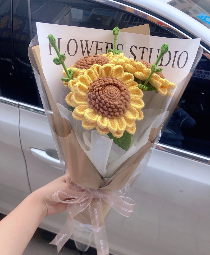 May include: A bouquet of crocheted sunflowers in a brown paper wrapper with a white ribbon tied around it. The bouquet is labeled "FLOWERS STUDIO".