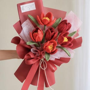 May include: A bouquet of crocheted red tulips with yellow centers and green leaves, wrapped in dark red and translucent white paper. A small white rectangular tag at the top features a floral illustration and the text A LITTLE FLOWER FOR YOU.